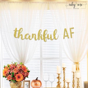 May include: Gold glitter Thanksgiving banner that reads "thankful AF". The banner is hanging in front of a window with white curtains. There is a table with a vase of orange and yellow flowers, a pumpkin that says "Give Thanks", and gold candle holders.