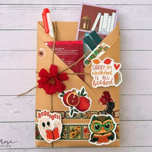 A fall-themed Blind Date with a Book bundle featuring a kraft paper envelope wrapped in twine, decorated with autumn stickers, and includes a highlighter, tea, book tabs, and a bookmark, adorned with a red flower, giving off cozy fall vibes.