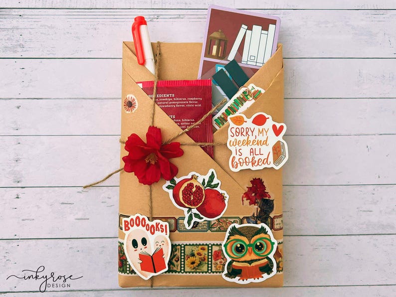 A fall-themed Blind Date with a Book bundle featuring a kraft paper envelope wrapped in twine, decorated with autumn stickers, and includes a highlighter, tea, book tabs, and a bookmark, adorned with a red flower, giving off cozy fall vibes.