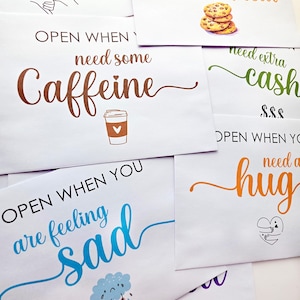 May include: A collection of white envelopes featuring colorful text and illustrations. Phrases include Open When You Need A Sweet Treat, Open When You Need Some Caffeine, Open When You Are Feeling Sad, and Open When You Need A Hug, with icons like cookies, a coffee cup, a rain cloud, and a heart.