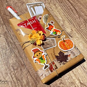 A fall-themed Blind Date with a Book bundle wrapped in kraft paper and twine, decorated with autumn stickers like a pumpkin , scarecrow, and includes a highlighter, tea, book tabs, and a bookmark, adorned with a flower, giving off cozy fall vibes.