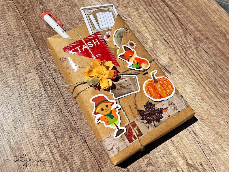 A fall-themed Blind Date with a Book bundle wrapped in kraft paper and twine, decorated with autumn stickers like a pumpkin , scarecrow, and includes a highlighter, tea, book tabs, and a bookmark, adorned with a flower, giving off cozy fall vibes.