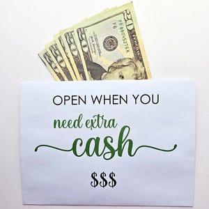May include: A white paper envelope containing several twenty-dollar bills. The envelope features the black text "OPEN WHEN YOU" followed by the green cursive text "need extra cash" and three black dollar signs at the bottom center.