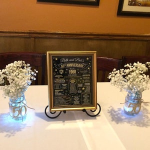 May include: A gold framed black chalkboard sign with gold lettering that reads "Beth and Bob's 50th Anniversary 1968" with a list of events that happened in 1968. The sign is on a black metal easel and is sitting on a white tablecloth. There are two mason jars with white flowers and gold glitter on the table.