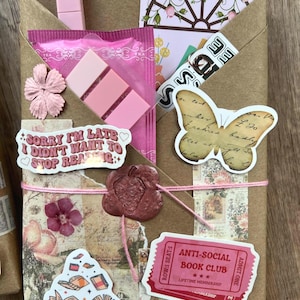 May include: A brown paper package tied with pink string, adorned with stickers, a pink clip, and a wax seal. Stickers include a butterfly, open books, and text that reads "Sorry I'm late I didn't want to stop reading." Also includes an "Anti-Social Book Club" ticket.