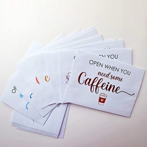 May include: A fan of white paper envelopes, each featuring black and colored text. The top envelope displays the phrase "OPEN WHEN YOU need some CAFFEINE" in black and dark red lettering, accompanied by a small illustration of a coffee cup.