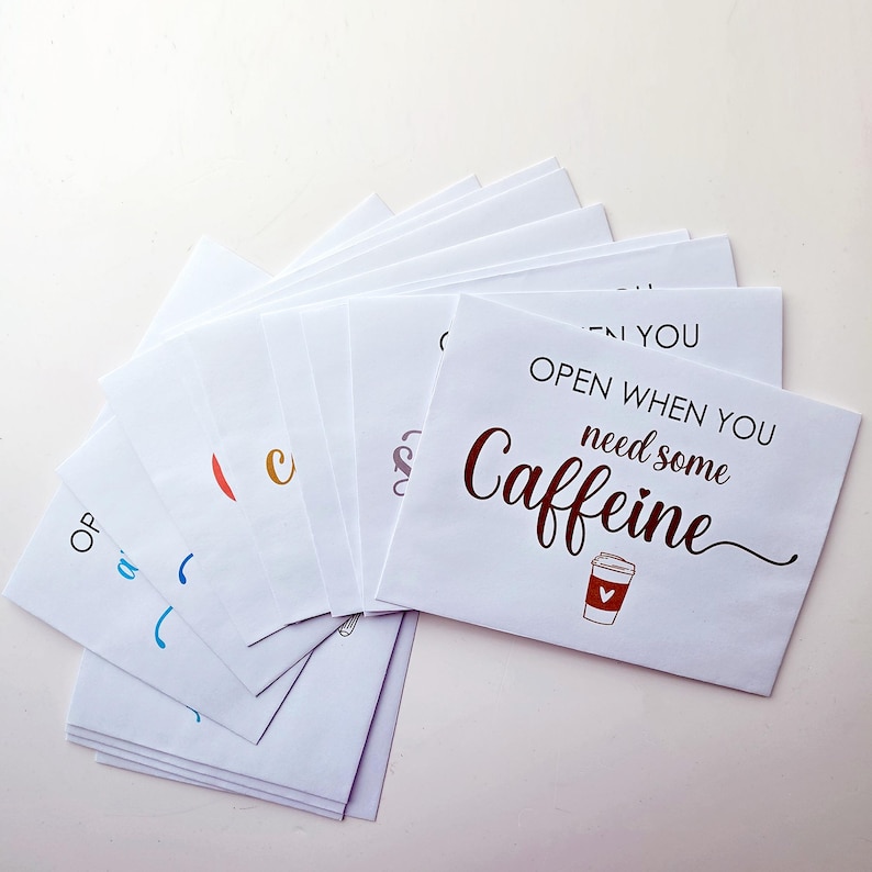 May include: A fan of white paper envelopes, each featuring black and colored text. The top envelope displays the phrase "OPEN WHEN YOU need some CAFFEINE" in black and dark red lettering, accompanied by a small illustration of a coffee cup.