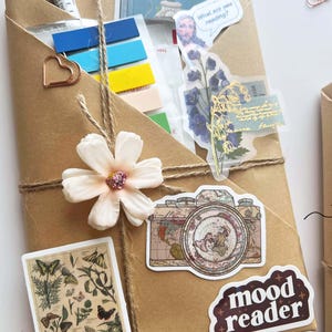 May include: A wrapped package in brown paper, adorned with stickers, a flower, and twine. Stickers include a vintage camera, butterflies, and the words "mood reader" and "What are you reading?" A pen and colorful sticky notes are visible.
