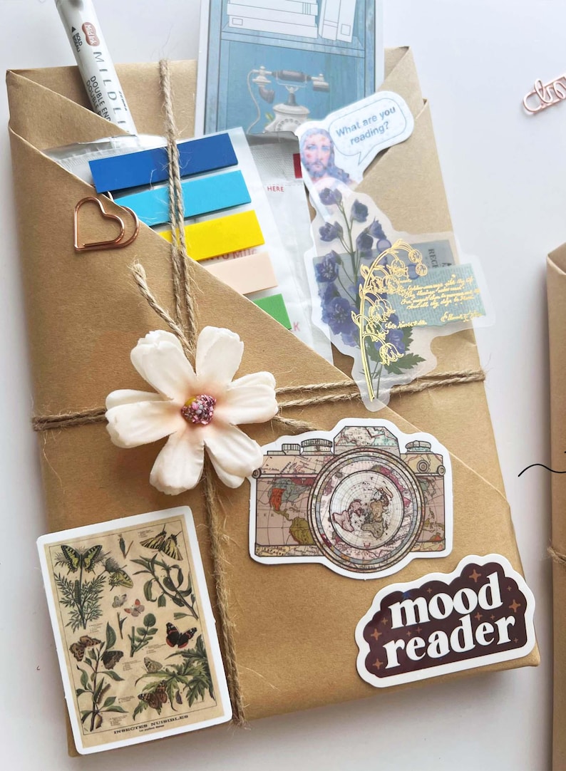 May include: A wrapped package in brown paper, adorned with stickers, a flower, and twine. Stickers include a vintage camera, butterflies, and the words "mood reader" and "What are you reading?" A pen and colorful sticky notes are visible.