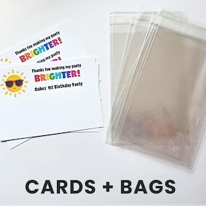 K&ouml;nnte beinhalten: Ein Stapel wei&szlig;er, rechteckiger Dankeskarten mit einer gelben Sonnengrafik mit Sonnenbrille und dem Text "Thanks for making my party BRIGHTER! Oakes' 1st Birthday Party" neben einem Stapel durchsichtiger, transparenter Kunststoffbeutel.