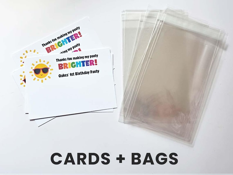 K&ouml;nnte beinhalten: Ein Stapel wei&szlig;er, rechteckiger Dankeskarten mit einer gelben Sonnengrafik mit Sonnenbrille und dem Text "Thanks for making my party BRIGHTER! Oakes' 1st Birthday Party" neben einem Stapel durchsichtiger, transparenter Kunststoffbeutel.