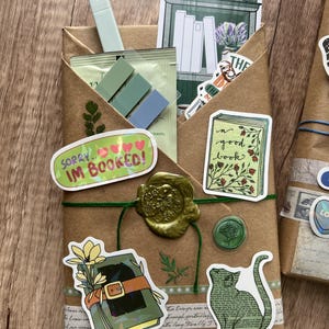 May include: A wrapped package with stickers and a wax seal. The package is brown with green twine and features stickers with book and cat illustrations, and the text "SORRY... IM BOOKED!". A light green highlighter and sticky notes are visible.