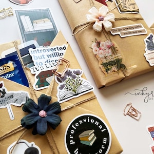May include: Wrapped gifts with decorative stickers and twine. The gifts are wrapped in brown paper and adorned with stickers featuring books, flowers, and inspirational quotes. A blue pen and tea bag are visible.