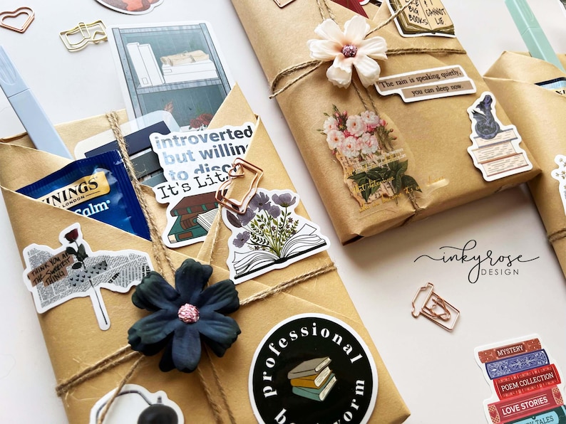 May include: Wrapped gifts with decorative stickers and twine. The gifts are wrapped in brown paper and adorned with stickers featuring books, flowers, and inspirational quotes. A blue pen and tea bag are visible.