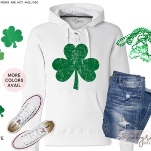 May include: White hoodie featuring a green shamrock graphic and the text "inkyrose design". The hoodie has a lace-up detail at the neck. The image also includes a pair of white trainers, blue jeans, and a green shamrock necklace.