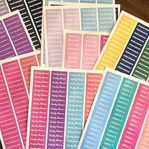 May include: Sheets of personalized name labels in various colors, including pink, purple, blue, and green. Each label features a name printed in a white, elegant font. The labels are arranged on a wooden surface.