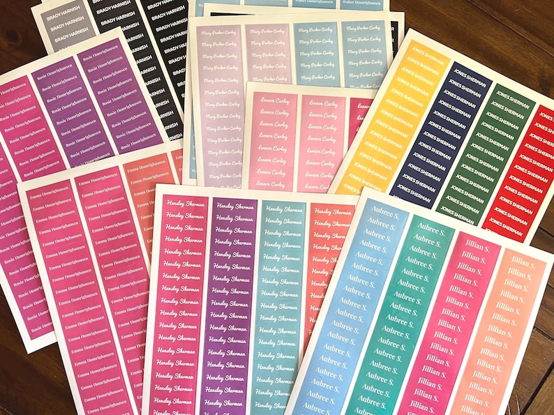 May include: Sheets of personalized name labels in various colors, including pink, purple, blue, and green. Each label features a name printed in a white, elegant font. The labels are arranged on a wooden surface.