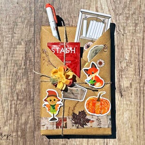 A fall-themed Blind Date with a Book bundle wrapped in kraft paper and twine, decorated with autumn stickers like a scarecrow and pumpkin, and includes a highlighter, tea, book tabs, and a bookmark, adorned with a flower, giving off cozy fall vibes.