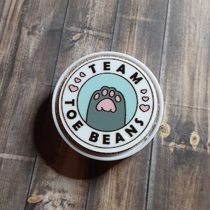 Peut inclure: Un autocollant rond blanc et scintillant avec les mots "TEAM TOE BEANS" encerclant un cercle bleu clair. À l'intérieur du cercle se trouve une patte de chat grise avec des coussinets roses. De petits coeurs roses sont entre les mots.