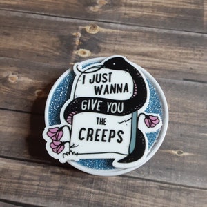 Puede incluir: Un pin redondo blanco con borde de purpurina azul. El pin presenta una serpiente negra enrollada alrededor de una lápida con el texto "I JUST WANNA GIVE YOU THE CREEPS" y flores rosas.