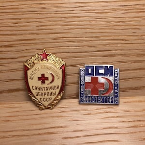 Può includere: Due spille smaltate vintage con croci rosse e mezzelune. La prima spilla è dorata con il testo "Народному Ополчению" e "Санитарной Обороны СССР". La seconda spilla è blu e argentata con il testo "Общественный Санитарный Инспектор" e "ОСИ".
