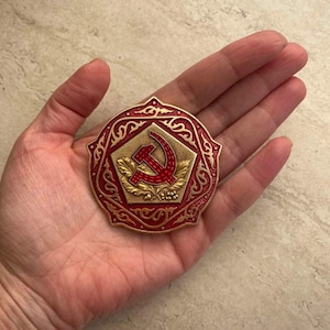 May include: A red and gold metal pin with a hammer and sickle design surrounded by an intricate floral pattern.