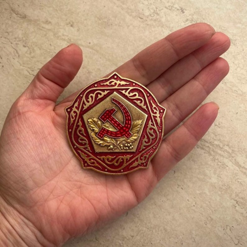 Puede incluir: Un pin de metal rojo y dorado con un dise&ntilde;o de martillo y hoz rodeado de un intrincado patr&oacute;n floral.