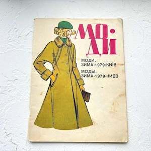 May include: Vintage fashion magazine cover featuring a woman in a long, olive green coat with a fur collar and a green beret. The magazine title is in large pink letters, with text in Russian below. The cover is slightly worn.