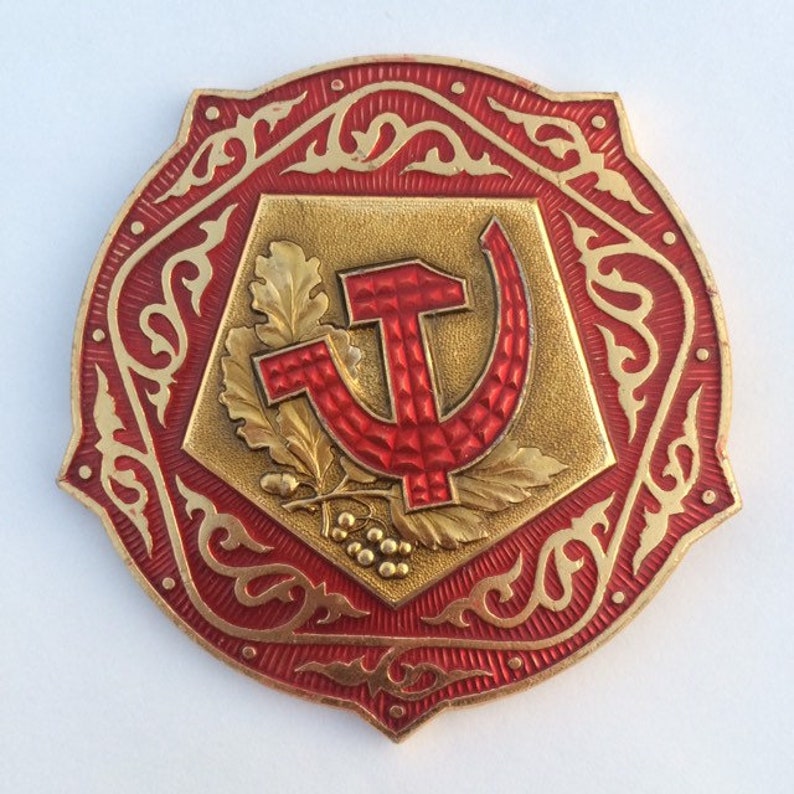 Puede incluir: Un pin de metal rojo y dorado con un dise&ntilde;o de martillo y hoz rodeado de un intrincado patr&oacute;n floral.