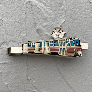 May include: A silver-toned metal tie clip featuring an enamel inlay of a tram. The tram design includes a red lower section, a cream middle, and blue window panels, topped with a silver pantograph. The tie clip has a polished metallic finish.