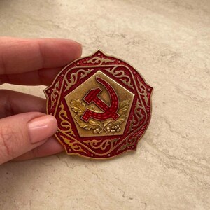 Puede incluir: Un pin de metal rojo y dorado con un dise&ntilde;o de martillo y hoz rodeado de un borde decorativo.