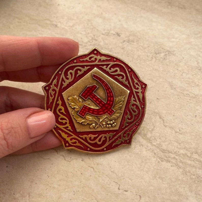 Puede incluir: Un pin de metal rojo y dorado con un dise&ntilde;o de martillo y hoz rodeado de un borde decorativo.
