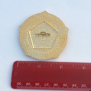 Puede incluir: Pin de metal dorado con una forma de pent&aacute;gono en el centro. El pin tiene un di&aacute;metro de 5 cm y una superficie texturizada.
