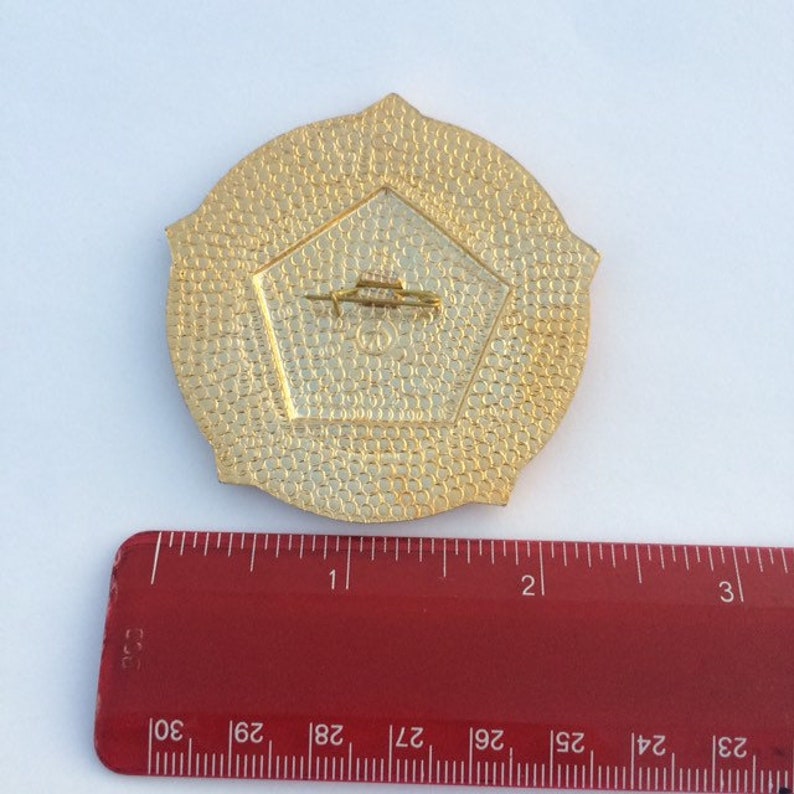 Puede incluir: Pin de metal dorado con una forma de pent&aacute;gono en el centro. El pin tiene un di&aacute;metro de 5 cm y una superficie texturizada.