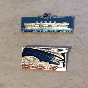 Pode incluir: Dois pinos de esmalte vintage que representam terminais de aeroporto. O pino superior é de prata com esmalte azul e apresenta um terminal de aeroporto estilizado com o texto "ЛЕНИНГРАД-АЭРОПОРТ ПУЛКОВО". O pino inferior é de prata com esmalte azul e vermelho e apresenta um terminal de aeroporto estilizado com um avião e o texto "КИЕВ-БОРИСПОЛЬ".