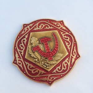 Puede incluir: Un pin de metal rojo y dorado con un dise&ntilde;o de martillo y hoz rodeado de un intrincado patr&oacute;n floral.