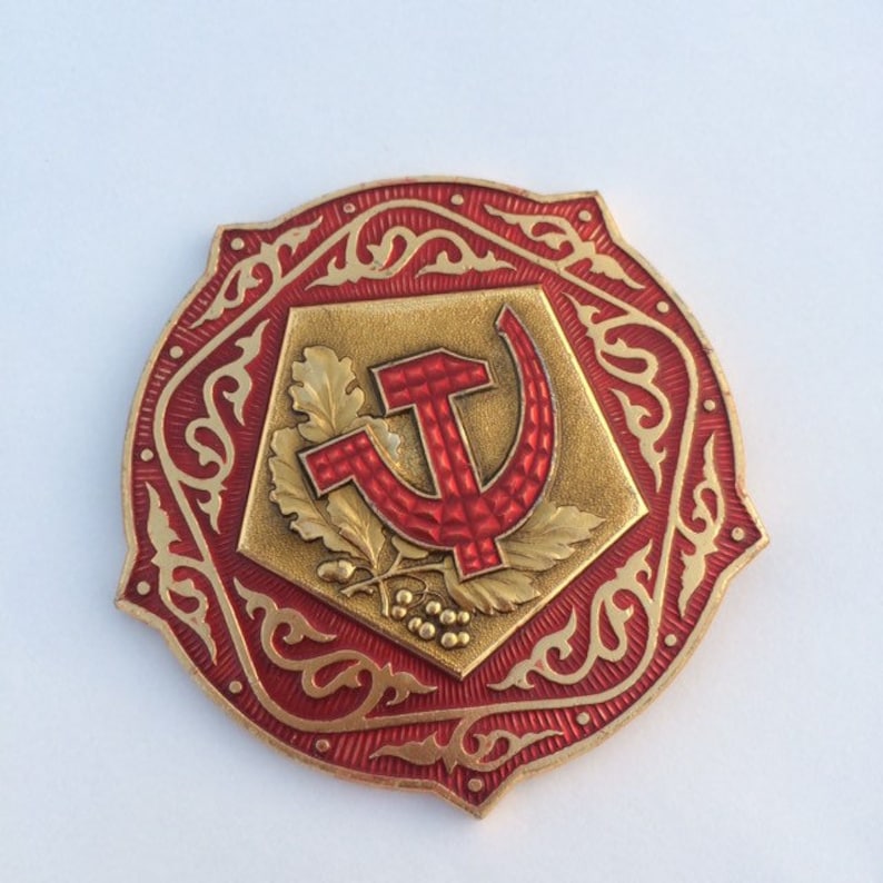 Puede incluir: Un pin de metal rojo y dorado con un dise&ntilde;o de martillo y hoz rodeado de un intrincado patr&oacute;n floral.