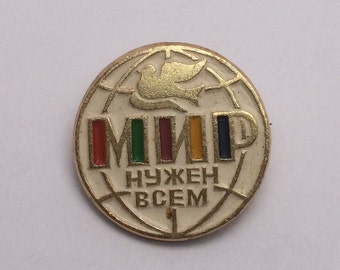 Puede incluir: Un broche circular de metal dorado con fondo de esmalte blanco y un patrón de cuadrícula de globo terráqueo. En la parte superior hay una paloma dorada. El texto cirílico dice "МИР" con barras verticales de colores y "НУЖЕН ВСЕМ" en oro.