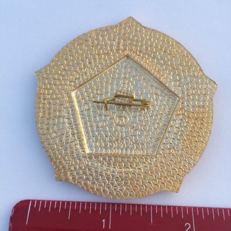 Puede incluir: La parte posterior de un pin de metal dorado con forma circular y superficie texturizada. El pin tiene un cierre de seguridad en la parte posterior.