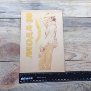 Könnte beinhalten: Ein Vintage-Magazin-Cover mit dem Text "МОДА 80" in Gelb. Die Illustration zeigt eine Frau in einem beigen Pullover, einer Hose und einem Hut. Am unteren Rand des Bildes befindet sich ein Lineal.