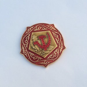 Puede incluir: Un pin de metal rojo y dorado con un dise&ntilde;o de martillo y hoz rodeado de un borde decorativo.