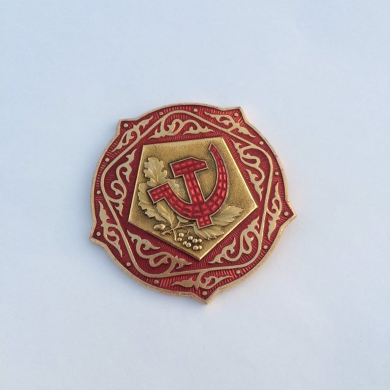 Puede incluir: Un pin de metal rojo y dorado con un dise&ntilde;o de martillo y hoz rodeado de un borde decorativo.