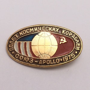 May include: A gold-colored oval pin with the text "ПОЛЕТ КОСМИЧЕСКИХ КОРАБЛЕЙ СОЮЗ - АПОЛЛО - 1975" around the edge. The center of the pin features a globe with the American and Soviet flags on either side.