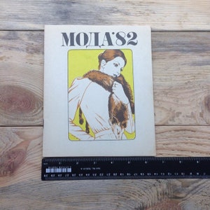 Könnte beinhalten: Ein Vintage-Magazincover mit einer Frau, die einen Pelzkragen und eine weiße Bluse trägt. Das Magazin trägt den Titel "MODA'82" in fetten, schwarzen Buchstaben.