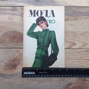 Könnte beinhalten: Vintage Modezeitschrift-Cover mit dem Wort "MODA" und der Zahl "80". Die Illustration zeigt eine Frau in einem grünen Outfit, Hut und Handtasche. Das Magazin liegt auf einem Holzgrund.