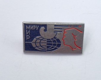 Puede incluir: Un pin de plata con un globo terráqueo azul y blanco, una silueta roja de una cabeza de persona y el texto "МИРУ ИР" en escritura cirílica.