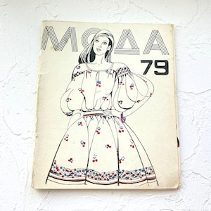 Op de afbeelding: Vintage mode tijdschriftcover met het woord "MODA" en het nummer "79". De illustratie toont een vrouw in een bloemenjurk met pofmouwen. De jurk heeft rode en blauwe bloemaccenten en een decoratieve rand.