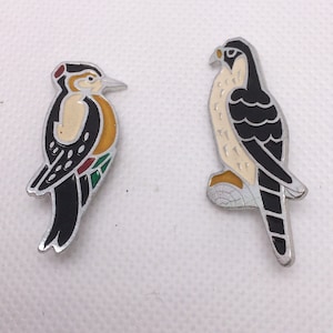 May include: Two enamel pins depicting a black and white woodpecker and a black and white falcon. The woodpecker is perched on a branch and the falcon is in flight. Both pins have a silver finish.