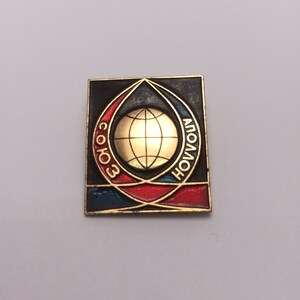 May include: A gold and black enamel pin with a globe in the center and the words "Soyuz" and "Apollo" written around the globe.