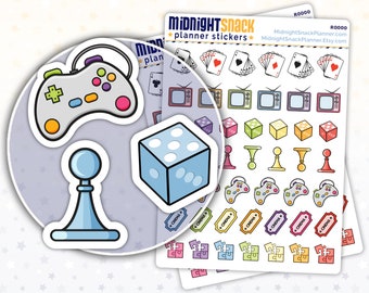 May include: A set of planner stickers featuring game-themed illustrations. The stickers include a game controller, a die, a game piece, playing cards, a television, and jigsaw pieces. The stickers are printed on white paper with a colourful design.